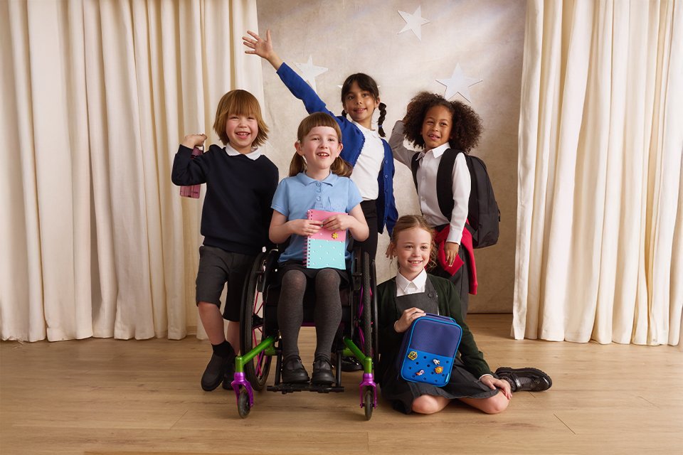 Five children in school uniforms pose on a stage with cream curtains and star decorations, one seated in a wheelchair holding a notebook, others standing and sitting with backpacks and lunch bags.