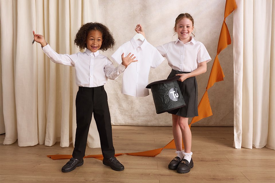Two children in white school shirts and dark uniform bottoms stand on a stage in front of cream curtains, holding a shirt on a hanger and a black TU-branded top hat, with an orange ribbon draped across the floor.