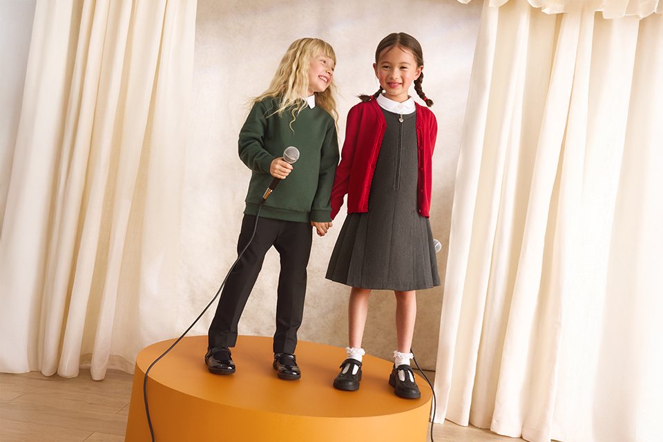 Two children in school uniforms standing on a round stage platform, holding microphones and performing in front of cream curtains, with a trailing microphone cable on the floor.