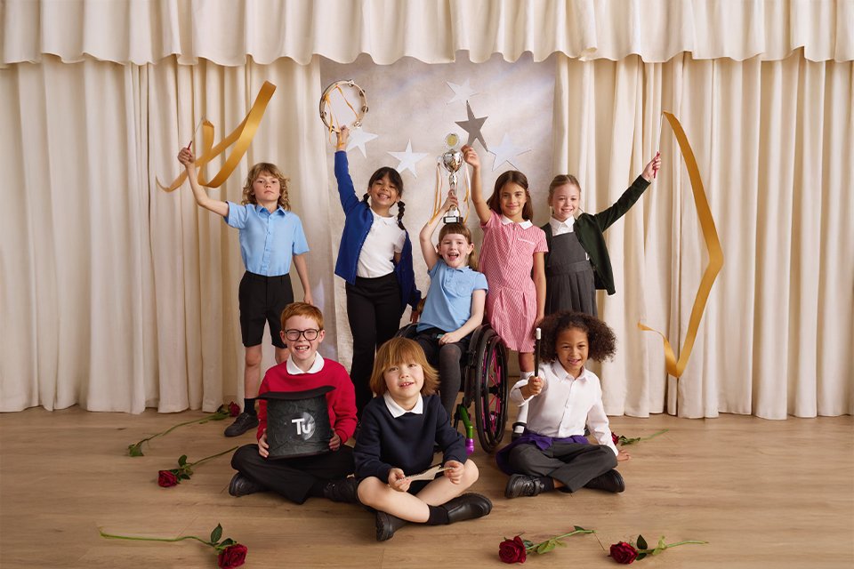 Children in school uniforms on a stage with beige curtains, holding ribbons, a trophy, and a tambourine, with star decorations and red roses on the floor.