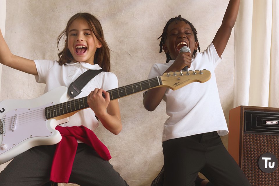 Two children in school uniforms kneeling on a stage, playing a white electric guitar and singing into a microphone next to a speaker with a TU logo.