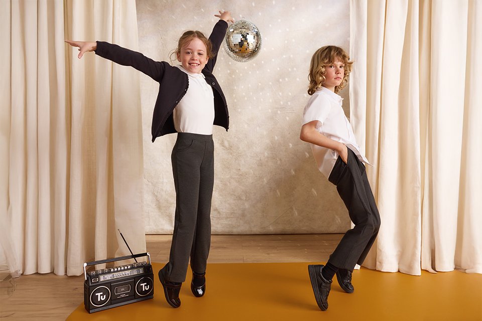 Two children in school uniforms dancing on a stage with cream curtains, one with outstretched arms and the other leaning back, with a disco ball and a TU-branded boombox on the floor.