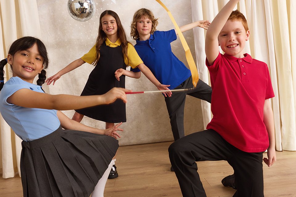 Four children in school uniforms on a stage with beige curtains, posing with rhythmic ribbons on sticks; a gold ribbon arcs through the air, with a silver disco ball and star decorations behind them.