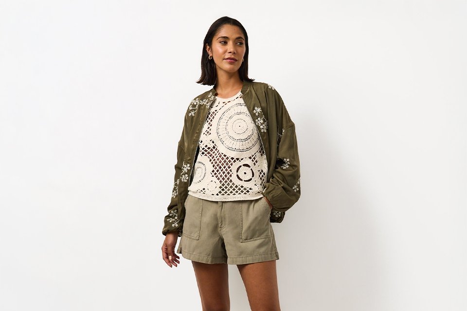 Woman wearing khaki shorts and a crochet top, with an embroidered khaki jacket and sandals.