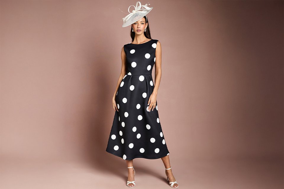 Woman wearing a black dress with white polka dots, a white fascinator and white shoes.