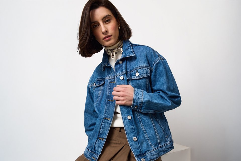 Mid-blue denim jacket styled over a white high-neck top with printed neck scarf, paired with brown tailored trousers for a smart casual everyday outfit.