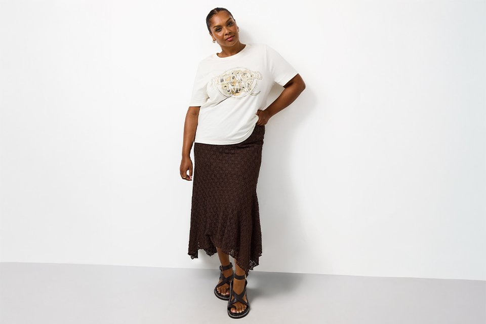 White graphic slogan t-shirt styled with a brown textured midi skirt and black strappy flat sandals, worn as a casual smart everyday outfit.