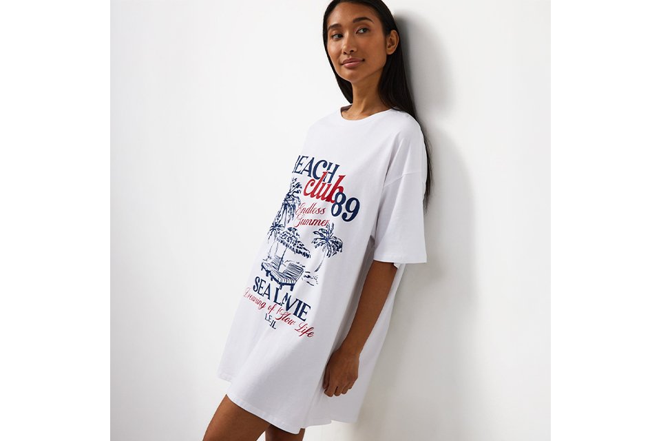 White oversized T-shirt nightdress featuring a Beach Club graphic print in blue and red with palm trees and seaside-inspired artwork.