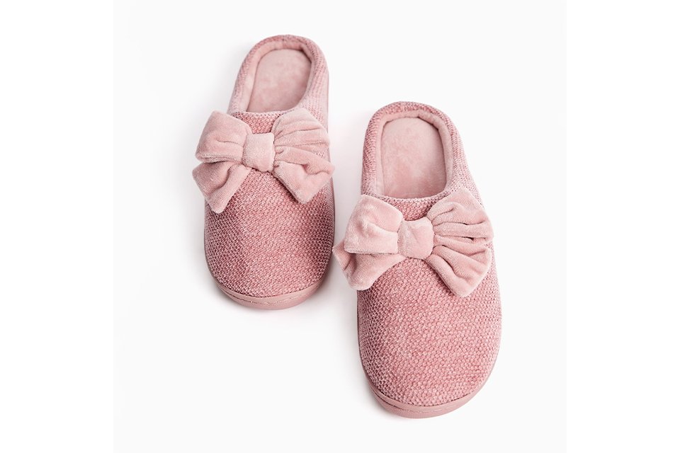 Pink textured mule slippers featuring oversized velvet bow detailing and a soft cushioned lining.