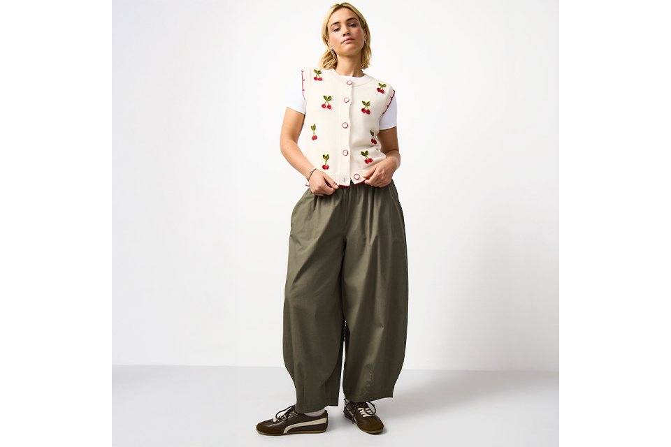 A woman wearing khaki balloon leg trousers, with a cherry print waistcoat.