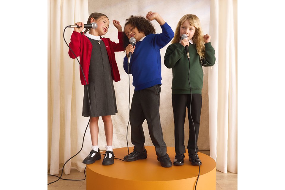 Three children standing on a stage platform singing into microphones, wearing colourful school uniform cardigans and jumpers during a school performance.