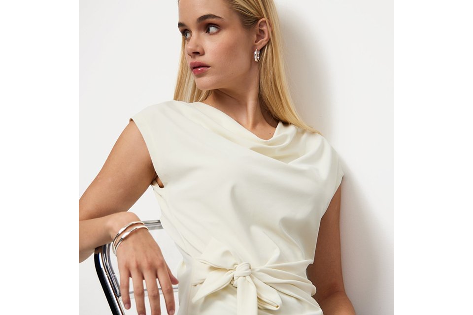 Ivory sleeveless blouse with a draped cowl neckline and side tie waist detail, styled with tailored trousers for an elegant contemporary look.
