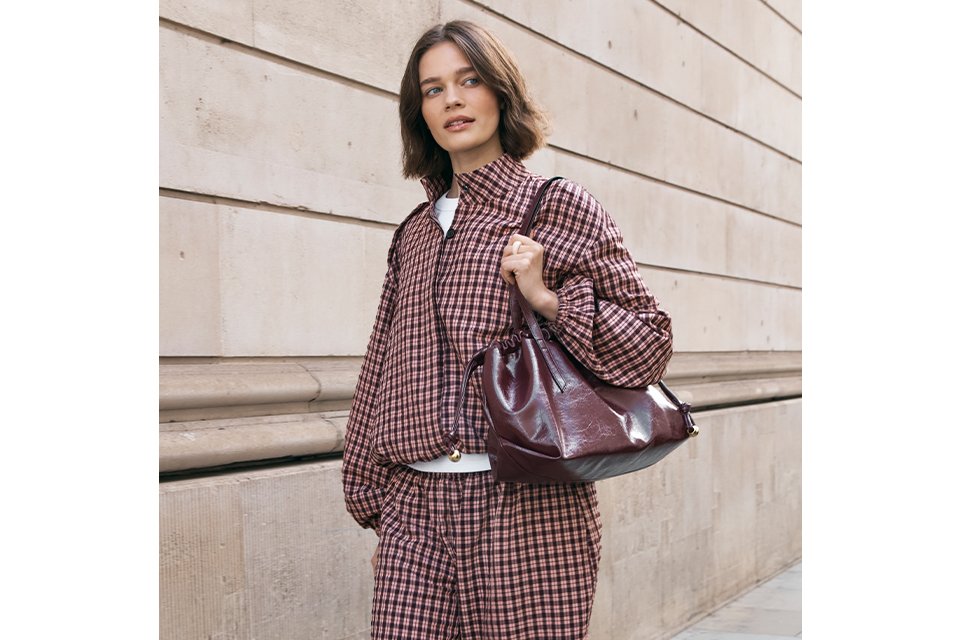 A woman wearing a matching co-ord of balloon style trousers, and matching jacket, in a red and pink check print, with black mary-jane style ballet pumps.