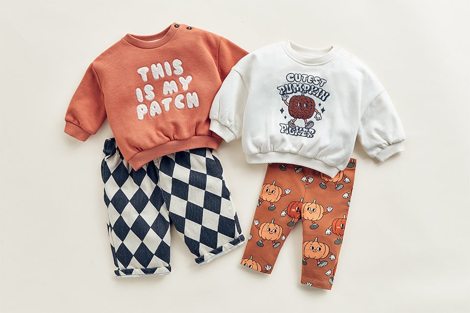 Baby Halloween outfit sets featuring pumpkin-themed sweatshirts paired with harlequin check trousers and pumpkin-print leggings, displayed in a flat-lay arrangement on a neutral background.