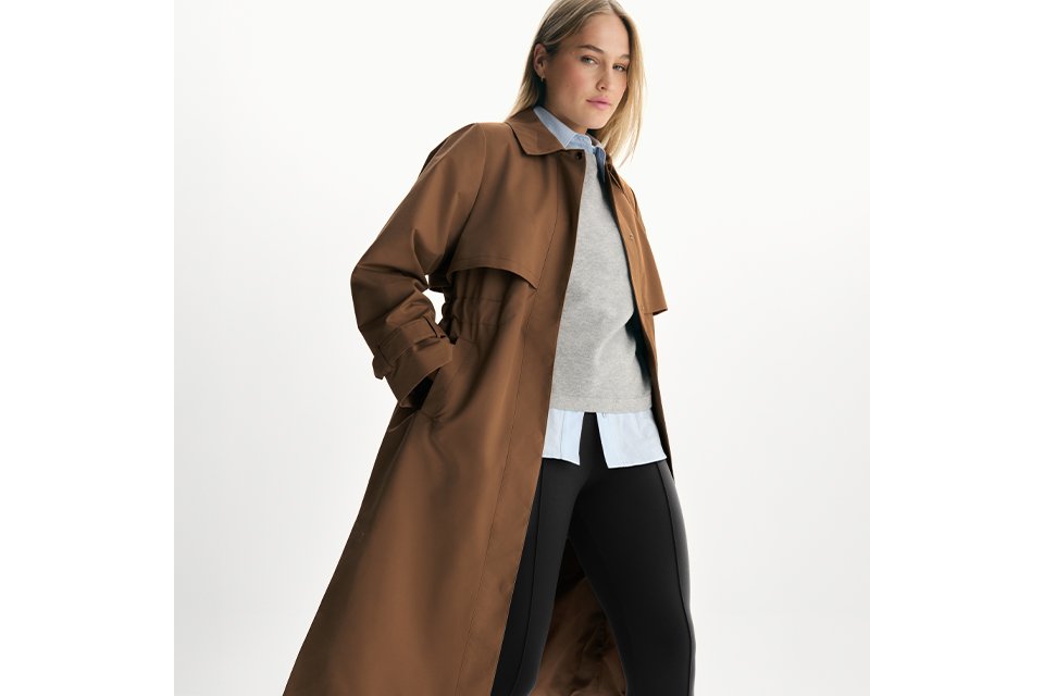 Brown trench coat outfit with grey sweater, blue shirt, black leggings, and black flats on a white background.