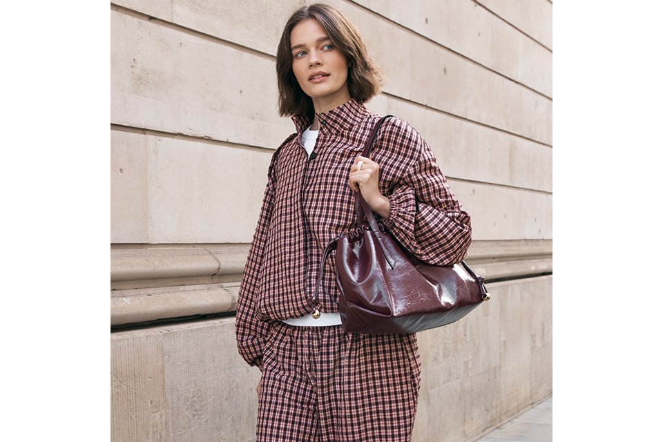 Person walking along a stone building wearing a matching burgundy and black checkered jacket and trousers set, styled with black Mary Jane flats and a burgundy leather shoulder bag.