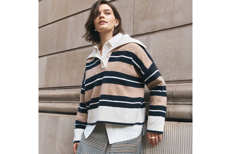 Person walking along a stone building wearing a matching burgundy and black checkered jacket and trousers set, styled with black Mary Jane flats and a burgundy leather shoulder bag.