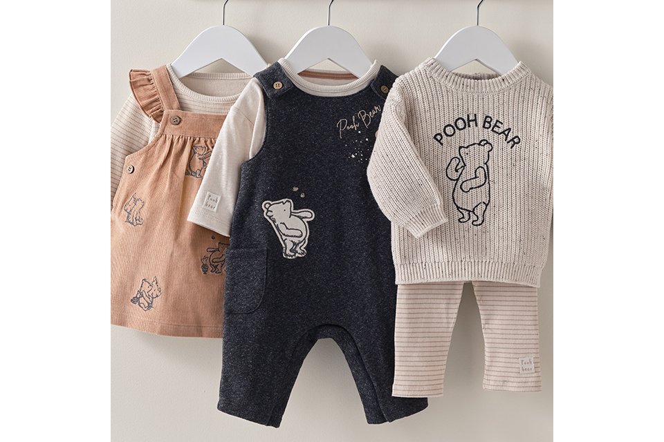 Disney Winnie the Pooh baby clothes collection featuring infant romper, knitted sweater, pinafore dress, and leggings in neutral beige, cream, and navy colours. Adorable newborn and baby outfit sets hanging on display.