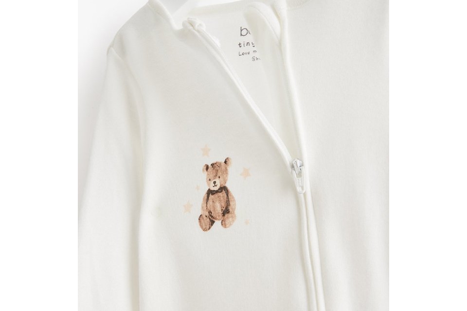 Cream baby zip-up sleepsuit featuring a teddy bear embroidery on the chest, made from soft cotton fabric and displayed on a hanger against a white background.