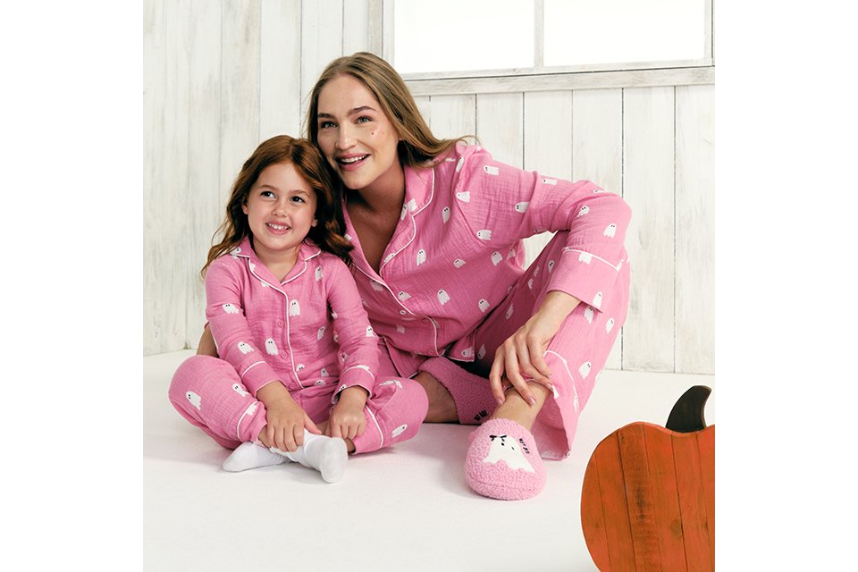 Person sitting on a bed wearing a cream graphic sweatshirt with a butter dish illustration and red gingham pyjama trousers, photographed in a cosy indoor setting.