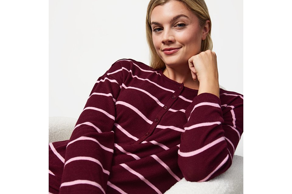Person seated in a white textured armchair wearing a burgundy ribbed lounge set with a long-sleeve crew-neck top and matching wide-leg trousers against a neutral studio background.