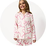 Child wearing a cream pyjama set with pink heart ribbon print, featuring a long-sleeve button-up sleep shirt and matching pajama trousers against a light studio background.