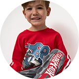 Child wearing a red Hot Wheels long-sleeve pyjama top with graphic print and grey checkered pajama bottoms featuring a Hot Wheels logo, seated on a bed against a light background.