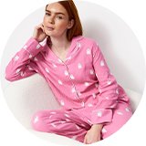 Person sitting in a white textured armchair wearing pink ghost-print pajama set with button-front shirt and matching lounge pants against a light studio background.