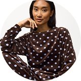 Person seated in a white textured armchair wearing a brown velour lounge set with white polka dots, featuring a long-sleeve top and matching relaxed-fit trousers against a light studio background.