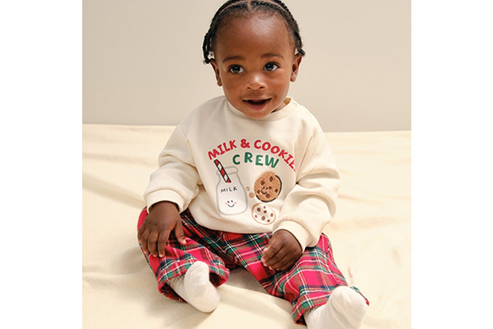 A baby wearing red tarten bottoms. with a cream jumper and a motif on it.