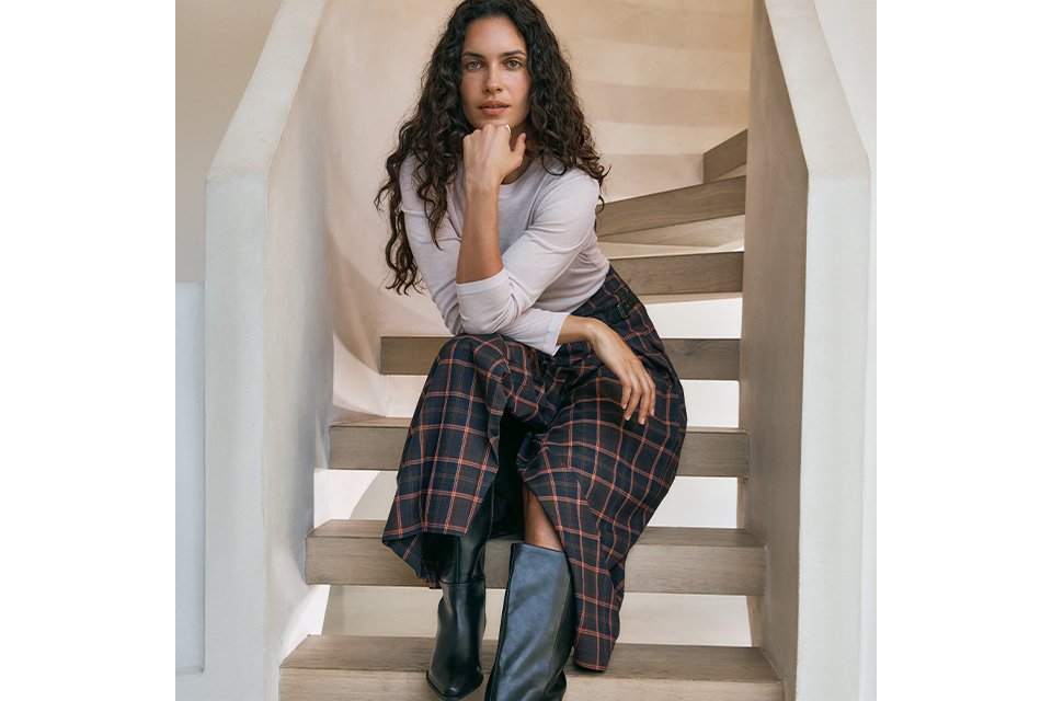 A woman wearing a neutral long sleeve top, with a black and red check skirt and black knee high boots.