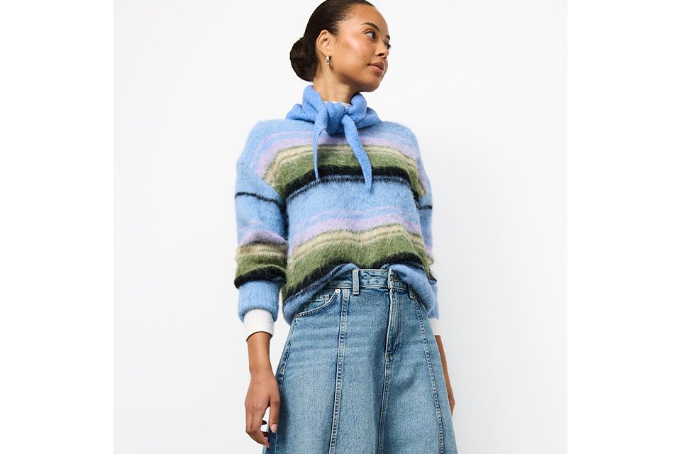 Light wash denim midi skirt styled with a soft blue striped knit sweater featuring a tie-neck detail, paired with cream ballet flats in a modern casual outfit.