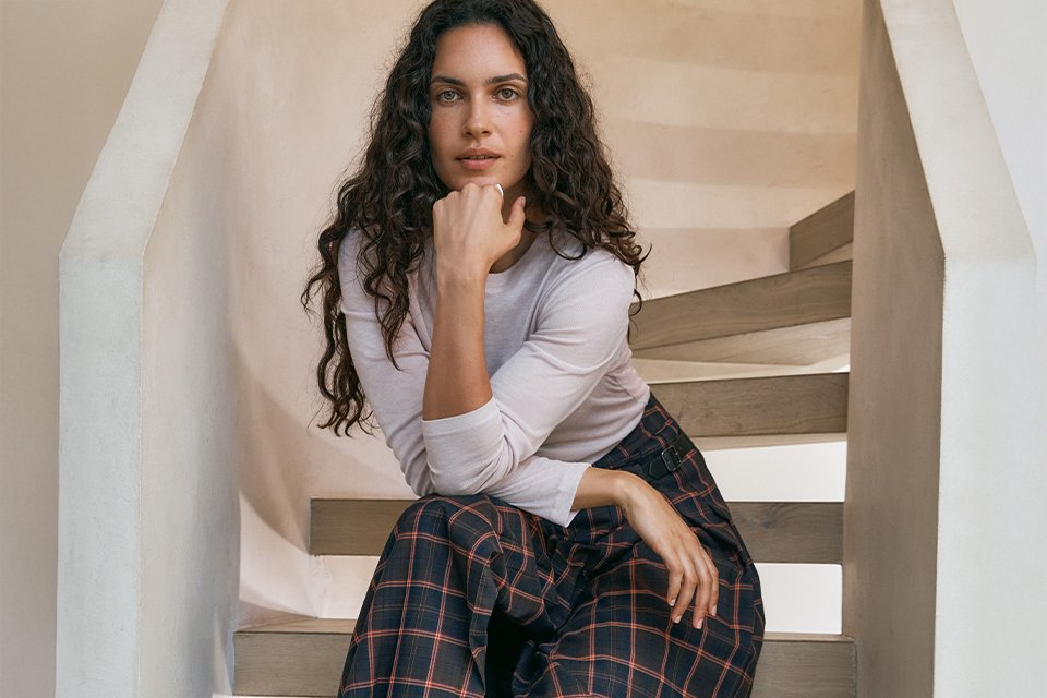 A woman wearing a neutral long sleeve top, with a black and red check skirt and black knee high boots