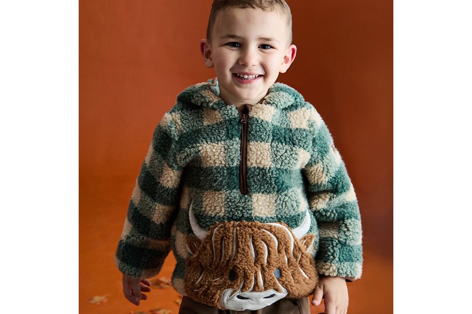 Close-up of a child wearing a brown graphic sweatshirt featuring playful dinosaur illustrations and bold cream lettering, photographed indoors against a neutral tiled background.
