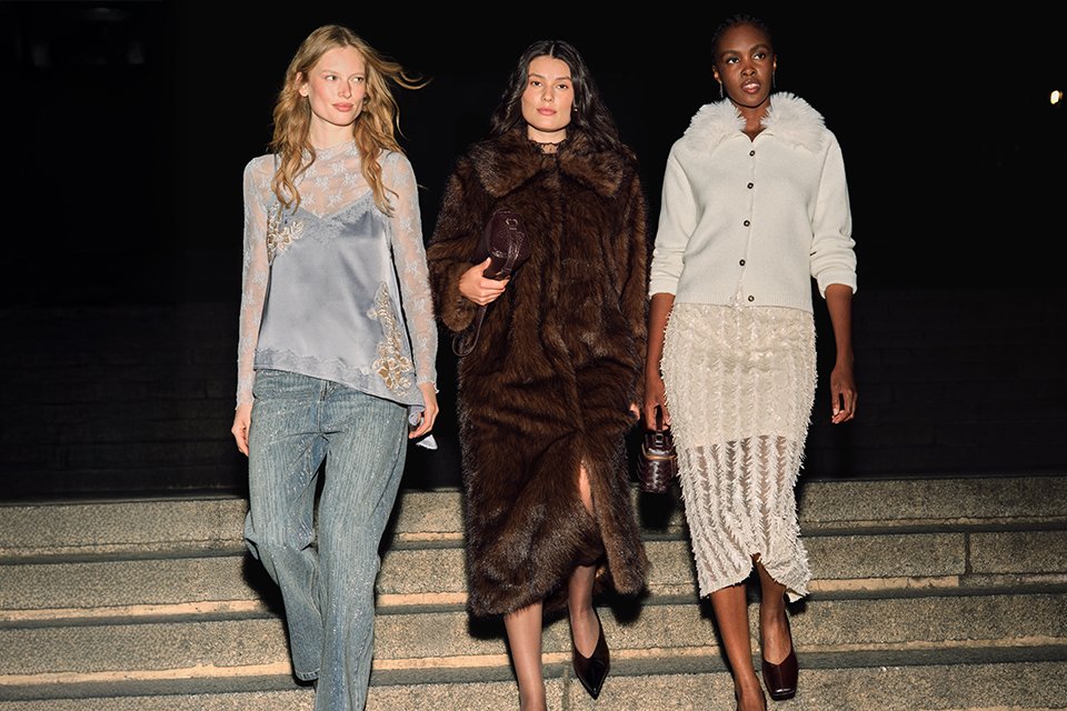 Three models showcasing luxury autumn-winter fashion on outdoor steps at night, featuring a lace-trim camisole with wide-leg trousers, a dark brown faux fur coat, and a textured knit cardigan paired with a fringed midi skirt.