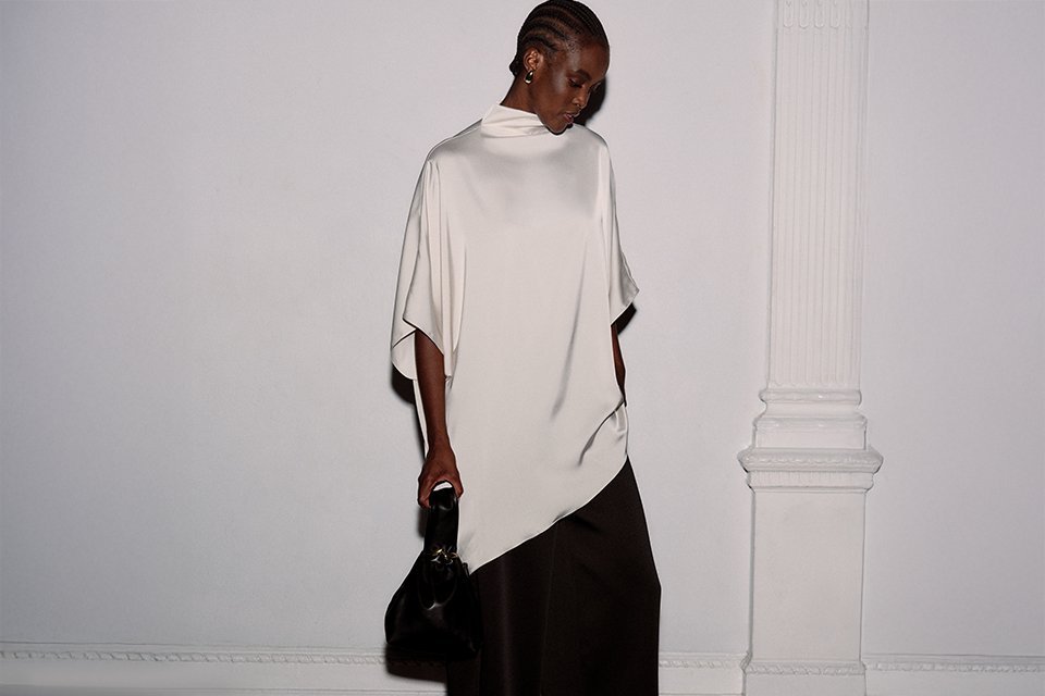 Women's cream draped high-neck top with asymmetric hem and cape-style sleeves, styled with wide-leg black trousers.