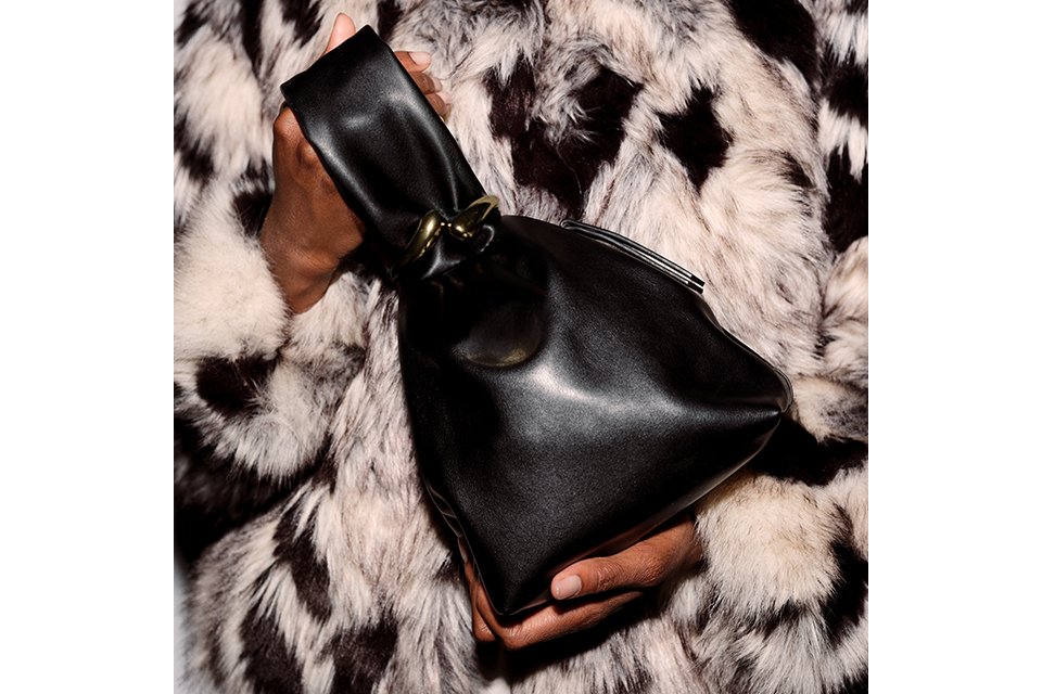 Women's black leather pouch clutch featuring a twisted handle and gold-tone detail, paired with a statement faux fur coat.