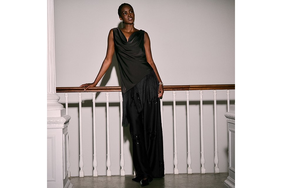 Women's black sleeveless draped top with tassel fringe hem and wide-leg black trousers, styled for eveningwear.