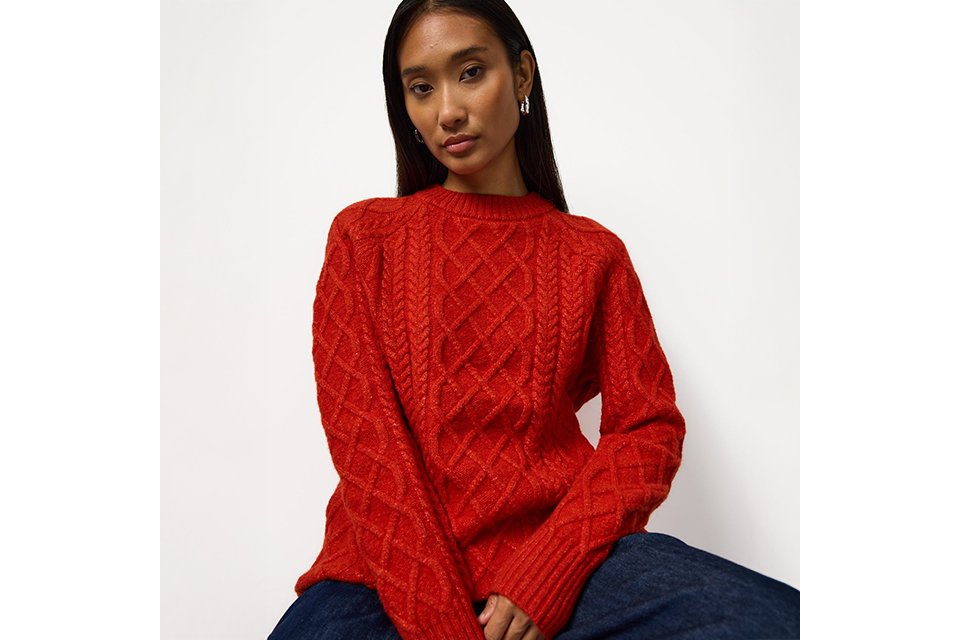 Bright red cable-knit crew neck sweater featuring an intricate textured pattern, styled with dark blue denim jeans for a classic casual winter fashion look.