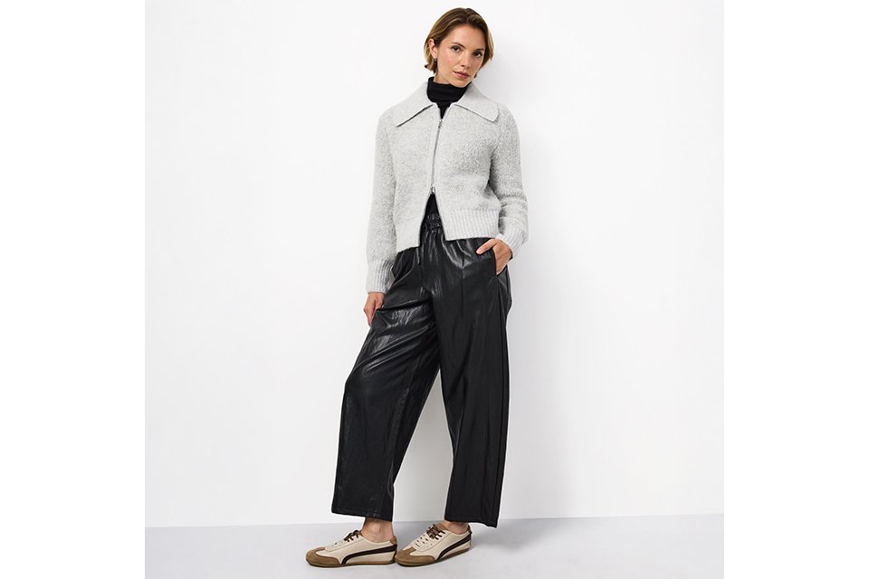 Light grey zip-front knit cardigan with a wide collar, layered over a black turtleneck and paired with black wide-leg faux leather trousers and casual low-top sneakers for a contemporary smart-casual look.