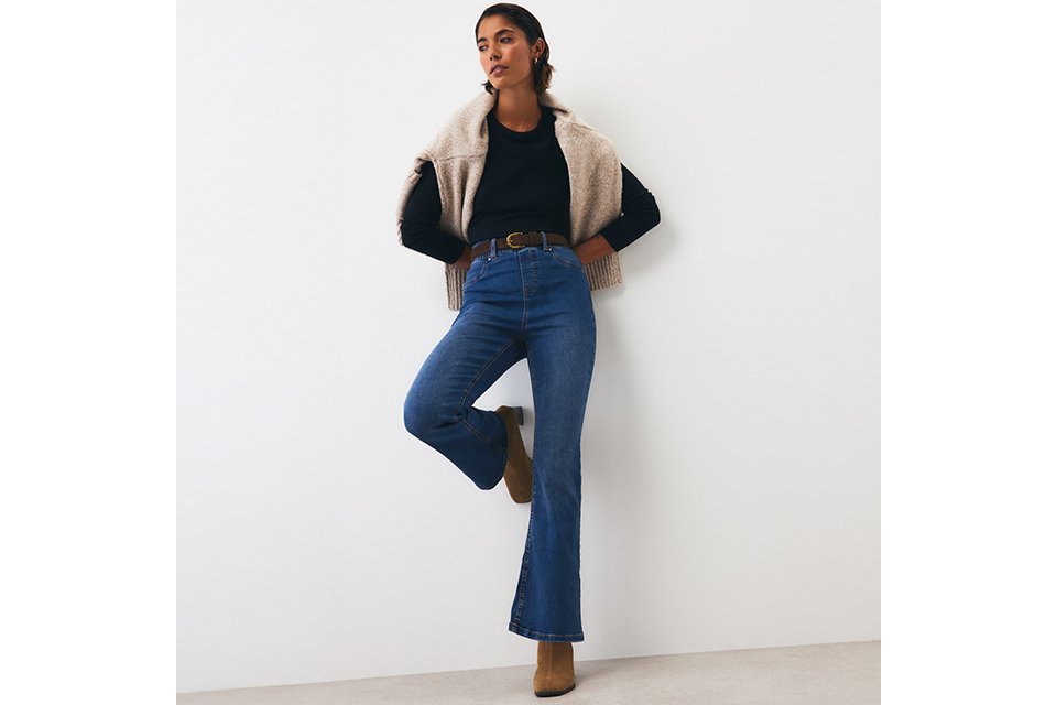 Blue high-waisted bootcut jeans paired with a black long-sleeve top, brown belt, ankle boots, and a beige knit sweater draped over the shoulders for a casual smart-casual look.