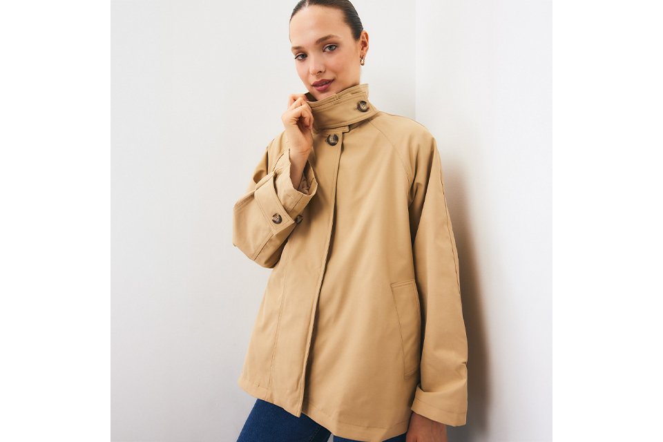 Beige high-collar utility jacket featuring oversized sleeves, button-detail cuffs, concealed front fastening, and patch pockets, styled with blue denim jeans in a minimalist studio setting.