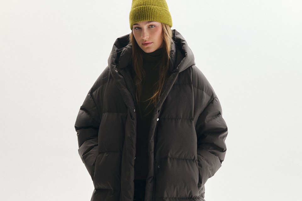 Long dark brown quilted puffer coat with an oversized hood, styled with a lime green knit beanie, black outfit, and chunky ankle boots for cold-weather winter fashion.