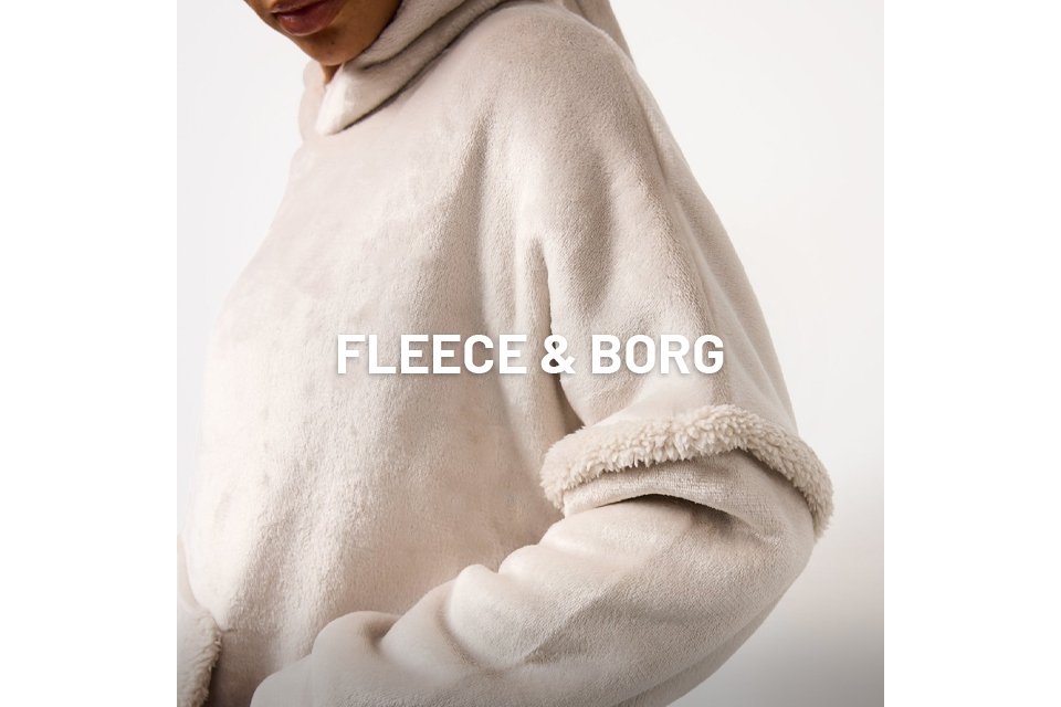 Close-up of a person wearing a light beige, plush fleece hoodie with a soft, textured finish. The garment features a cosy hood and a thick, sherpa-like trim detail on the sleeve. The person’s hand is partially tucked into a front pocket.