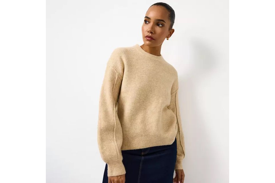 Person wearing a beige ribbed knit sweater paired with a dark skirt, standing against a plain white background.