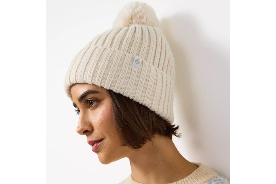 Close-up of a person wearing a cream-colored knitted winter hat with a pom-pom on top, paired with a light beige sweater featuring a textured pattern.