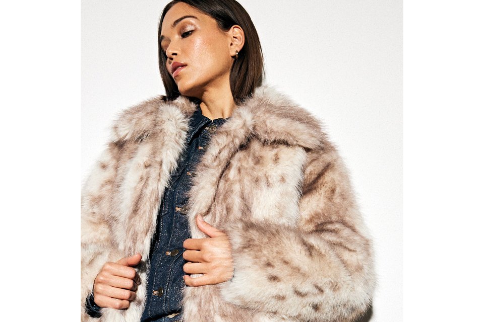 Person dressed in a thick, light brown and cream faux fur coat layered over a dark textured top, shown from the waist up.