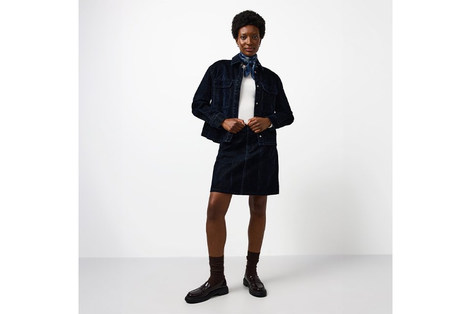 Person wearing a dark denim jacket and matching denim skirt, styled with a white top and black ankle boots, standing against a minimal white backdrop.