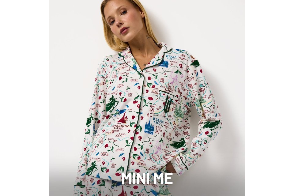 Person wearing a white pajama set with colorful illustrated prints, including castles, trees, and text. The top is button-up with dark piping along the edges, and the pants have a matching pattern. The background is plain white.