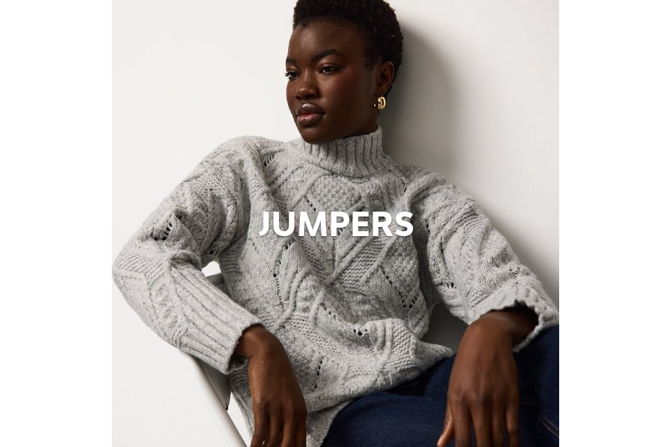 Person seated on a white chair wearing a light gray cable-knit sweater with a high neckline and dark blue jeans. 